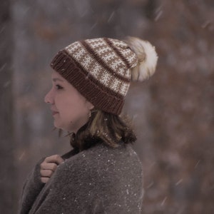 May include: A woman wearing a brown and white knit hat with a white pom pom. She is wearing a brown sweater and is standing in the snow.