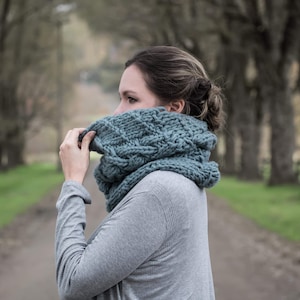 May include: A woman wearing a gray sweater and a chunky blue knitted cowl neck scarf. The scarf is made with a cable knit pattern.
