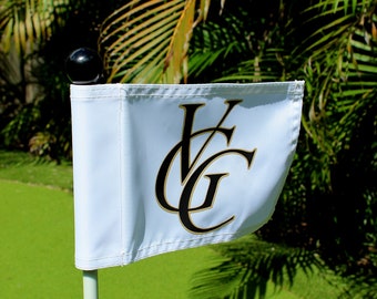 Può includere: Una bandiera da golf bianca con le lettere "VG" in nero e oro, su un green. La bandiera è su un palo bianco, sormontato da una palla nera, con palme sullo sfondo.