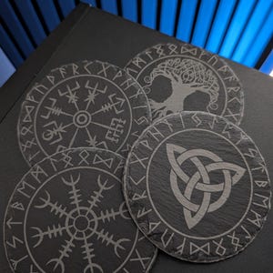 Könnte beinhalten: Drei runde Untersetzer aus schwarzem Schiefer mit eingravierten Motiven. Einer zeigt ein Vegvisir-Symbol, einer einen Triquetra-Knoten und einer einen Lebensbaum. Jeder Untersetzer ist von einem Ring aus in den dunklen Stein gravierten Runen umgeben.