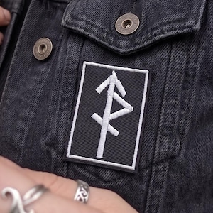 May include: A black denim vest featuring a rectangular embroidered patch on the chest. The patch has a black background with a white border and a white runic symbol stitched in the center. The vest has metal buttons and a classic collar design.