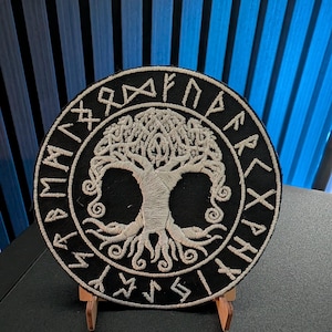 Könnte beinhalten: Ein kreisförmiger, schwarz bestickter Aufnäher mit einem weißen, gestickten Lebensbaum-Motiv in der Mitte. Der Baum ist von einem Rand mit weißen, gestickten Runenzeichen umgeben. Der Aufnäher steht auf einem kleinen Holzständer.