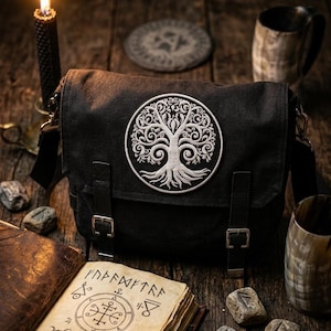 Könnte beinhalten: Eine schwarze Umhängetasche mit einem großen, kreisförmigen, weiß gestickten Baummotiv auf der Frontklappe. Die Tasche verfügt über zwei vertikale Riemen mit silberfarbenen Metallschnallen zum Verschließen und einen verstellbaren Schultergurt.