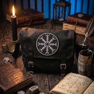 Op de afbeelding: Een zwarte schoudertas met een cirkelvormig, zilverkleurig Vegvisir-symbool op de voorflap. De tas is voorzien van twee verticale riemen met metalen gespen voor de sluiting en een verstelbare schouderriem bevestigd met metalen hardware.