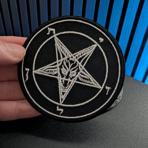 May include: A circular black embroidered patch featuring a white stitched pentagram design. The center of the star contains a detailed goat head illustration, and the entire emblem is enclosed within a thin white circular border.