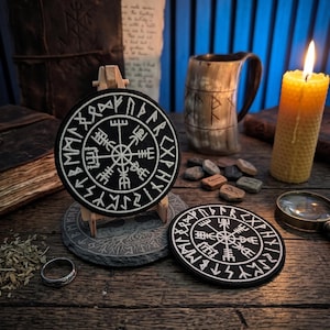 May include: Two circular black patches featuring white embroidered Vegvisir symbols surrounded by runic inscriptions. One patch is displayed upright on a small wooden easel, while the other lies flat on a dark wooden surface next to a horn mug.