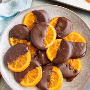 May include: A plate of chocolate-covered orange slices. The orange slices are cut into rounds and dipped in dark chocolate, with a sprinkle of sea salt on top. The image is a close-up shot.