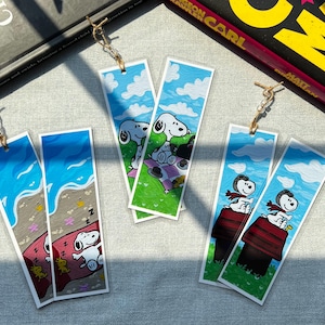 Peut inclure: Six marque-pages rectangulaires en papier illustrés avec le personnage Snoopy. Deux montrent Snoopy dormant sur une serviette de plage, deux sur une nappe de pique-nique et deux sur une niche. Chaque marque-page possède une boucle en fil doré.