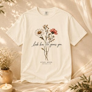 May include: A cream-colored short-sleeve t-shirt featuring a graphic of wildflowers and the text "look how He grows you. EVEN HERE. MATTHEW 6:28" printed in black ink on the center of the chest. The shirt has a crew neckline and a standard fit.
