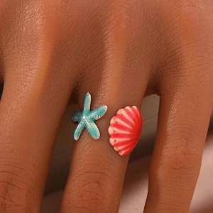 May include: A ring with a turquoise starfish and a red seashell design. The ring is open, allowing for an adjustable fit. The ring is a beach-themed accessory.