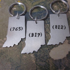 May include: Three silver keychains shaped like the state of Indiana with the area codes (765), (317), and (812) engraved on them.