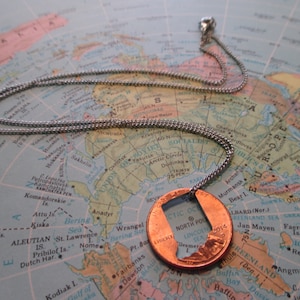 May include: A silver chain necklace with a copper penny pendant. The penny has a cutout of the state of Alaska, with the words "North Pole" and "Lincoln" visible. The necklace is displayed on a world map.