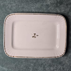 May include: A rectangular white ceramic serving platter with rounded corners and a fluted edge. The rim is accented with a thin gold-colored line, and a small, dark, stylized floral motif is centered on the flat surface of the dish.