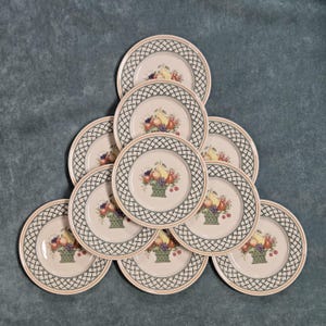 May include: A set of nine round ceramic plates arranged in a pyramid formation. Each plate features a cream-colored center with a central illustration of a fruit basket containing pears, grapes, and cherries, surrounded by a black lattice border.