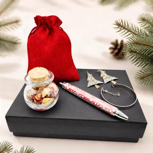 May include: A black rectangular gift box containing a red drawstring pouch, a small glass jar with a cork stopper filled with dried flower petals, a red and white snowflake patterned pen, two silver-toned Christmas tree earrings, and a silver bracelet.