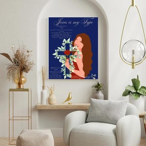May include: A framed art print with the text "Jesus is my Type" on a navy blue background. The artwork features a woman with long brown hair, a cross, and floral elements. The print is displayed in a modern living room setting.