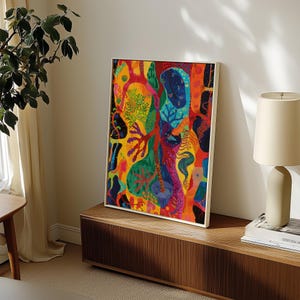 May include: Abstract art print in a white frame. The artwork displays a vibrant mix of colours, including red, yellow, green, and blue, with organic shapes and patterns. The painting is displayed on a wooden cabinet, with a lamp and books nearby.