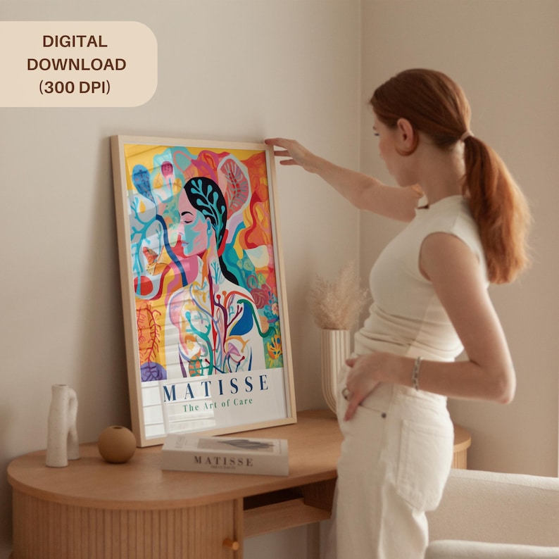 May include: A framed art print featuring a colorful abstract portrait with the text "MATISSE The Art of Care." The print has a digital download label. The artwork is displayed on a wooden table.