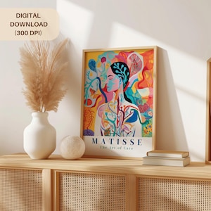 May include: A framed art print featuring a colorful illustration of a person with a floral design. The print has the text "MATISSE The Art of Care" and is displayed on a wooden cabinet with a vase of pampas grass and a decorative ball.