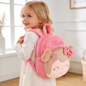 Op de afbeelding: Roze pluche rugzak met een cartoon meisjesgezicht ontwerp. De rugzak heeft de naam "Olivia" in glitterletters. Het gezicht van het meisje heeft een strik, een bloem en een glimlachende uitdrukking. De rugzak heeft een handvat aan de bovenkant en verstelbare bandjes.