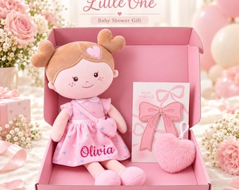 May include: A plush doll with brown hair styled in heart-shaped buns, wearing a pink dress with blue hearts and the name Olivia printed on it. The doll is inside a pink gift box with a Best Wishes card featuring a pink bow and a fuzzy heart keychain.