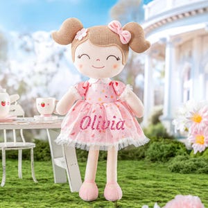 May include: A plush doll with brown hair styled in two high pigtails, adorned with a pink bow and flowers. The doll wears a pink dress with a strawberry pattern and the name "Olivia" in glitter. The doll is standing on green grass.