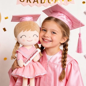 May include: A young girl in a pink graduation gown and cap smiles, holding a plush doll also wearing a graduation cap and gown. The doll holds a banner that reads "Congratulations". A banner that says "GRADUATE" is in the background.