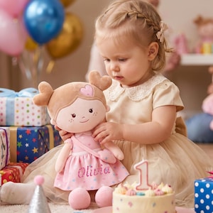 Può includere: Una bambola di peluche con un viso a forma di cuore e un vestito rosa con il nome "Olivia" ricamato. La bambola è tenuta in braccio da una bambina. Una torta di compleanno con una candela a forma di numero 1 è in primo piano, insieme a regali confezionati e palloncini.