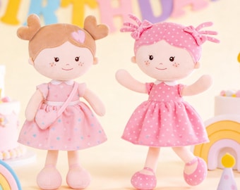 Puede incluir: Dos muñecas de peluche con mejillas rosadas y caras sonrientes. Una muñeca tiene el pelo castaño en coletas con detalles de corazón, con un vestido rosa con detalles de corazón y un bolso a juego. La otra muñeca tiene el pelo rosa y un vestido rosa de lunares.