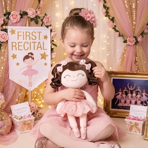 May include: A young girl in a pink dress holds a ballerina doll with brown hair and a pink tutu. The doll matches the theme of the "First Recital" banner in the background. The scene is decorated with roses and gold accents.