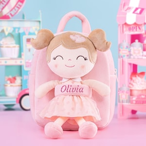 May include: A plush doll with a smiling face, wearing a pink dress and a matching backpack. The doll has brown hair in pigtails and the name "Olivia" embroidered on the dress. The backpack has a top handle.