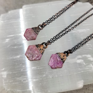 May include: Three necklaces with pink and copper pendants. Each pendant is a raw, rough-cut pink crystal with a copper backing. The necklaces are on a black chain.