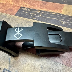 May include: A matte black belt buckle featuring a rectangular base and a hinged mechanism. The top surface displays a white graphic symbol resembling a stylized brand or mark. The buckle has a smooth finish and a mechanical design with a lever.