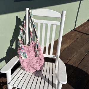 May include: A pink crocheted tote bag with floral patterned straps and accents hangs on a white wooden rocking chair. The bag has a textured appearance and is positioned against a light green wall and wooden deck.
