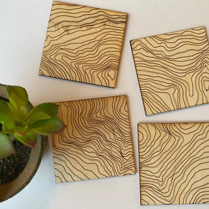 May include: Four square wooden coasters with a brown wood grain and a black topographic map design.
