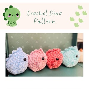 May include: Four crocheted dinosaur plushies in different colors: light blue, pink, red, and turquoise. The plushies are sitting on a green surface. Crochet Dino Pattern.