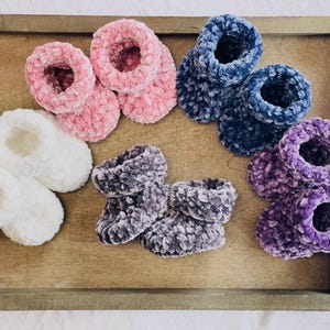 May include: A collection of baby booties in various colors, including pink, white, blue, gray, and purple. The booties are made of a soft, plush material and are arranged on a wooden tray. The booties are designed for infants.