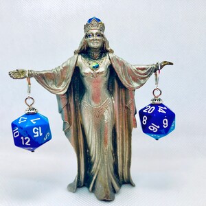 May include: A silver figurine of a woman wearing a crown and a blue dress, holding two blue and silver dice earrings. The dice have white numbers on them.