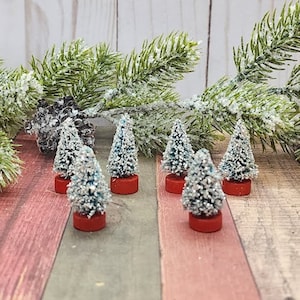 May include: Miniature Christmas trees with silver glitter and red bases are arranged on a wooden surface. Green pine branches with snow-like accents and a pine cone are in the background. Festive holiday decor.