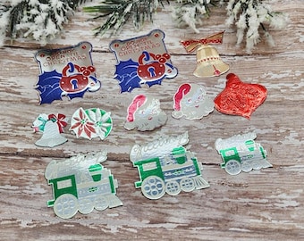 May include: A collection of vintage Christmas-themed die-cut decorations. The assortment includes ornaments shaped like bells, trains, Santa hats, and tags with the text "DO NOT OPEN TIL CHRISTMAS". The color palette features red, green, silver, and gold.