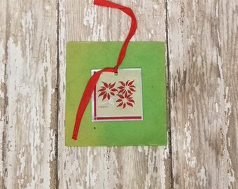 May include: A green card with a silver tag attached. The tag has a red ribbon tied around it and features a design of red poinsettia flowers. The tag says "Seasons Greetings".