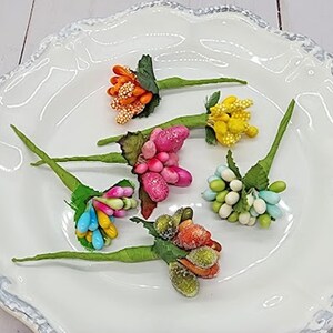 May include: A collection of colorful artificial flower stems with various shades of orange, pink, yellow, blue, and green. The stems are arranged on a white, scalloped-edge plate, showcasing the vibrant hues of the floral accents.