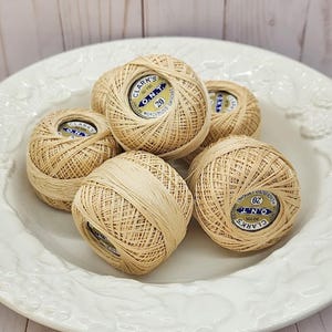 May include: A collection of six beige mercerized cotton crochet thread balls, each featuring a circular label with the text Clark's O.N.T. 20, arranged in a cluster. The thread is wound into compact spheres, showcasing a consistent light tan color.
