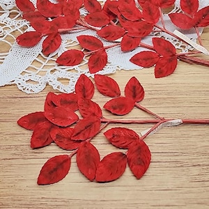 May include: Several red artificial leaf stems with three leaves each. The leaves have a textured appearance and are arranged on thin, brown stems. The background includes a white lace doily and a wooden surface.