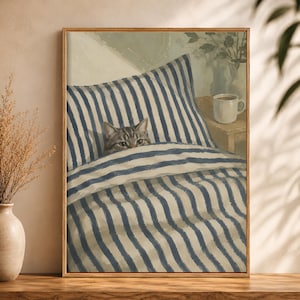 May include: A framed print of a cat peeking out from under a blue and white striped blanket and pillow. A mug of coffee sits on a small wooden table next to the bed. The artwork has a warm, inviting feel.
