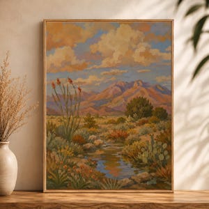 May include: A framed landscape painting depicts a desert scene with mountains under a cloudy sky. The artwork features a stream, cacti, and colorful vegetation in shades of orange, green, and blue. The frame is a light brown color.