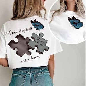 Puede incluir: Camiseta blanca con una mariposa azul y una pieza de rompecabezas gris con el texto "A piece of my heart lives in heaven" y "Allison 1996-2022".