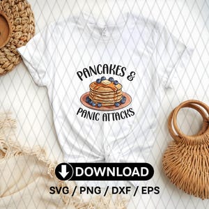 Può includere: Maglietta bianca con una grafica di una pila di pancake con mirtilli e sciroppo, e il testo "Pancakes & Panic Attacks".