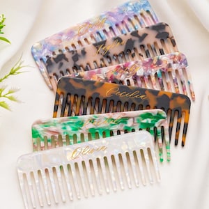 May include: A collection of personalized hair combs in various colors and patterns, including tortoiseshell, green, and white. Each comb has a name in gold script. The combs are arranged on a white fabric surface.