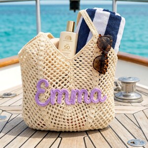 K&ouml;nnte beinhalten: Beige geh&auml;kelte Tragetasche mit dem Namen "Emma" in lila Garn. Die Tasche enth&auml;lt eine Sonnencreme, ein gestreiftes Handtuch und eine Sonnenbrille. Die Tasche befindet sich auf einer Holzoberfl&auml;che mit t&uuml;rkisfarbenem Meer im Hintergrund.